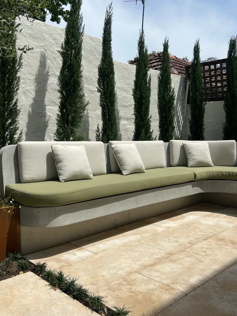 Pale built-in seating, olive cushions and slender upright plants against a light wall